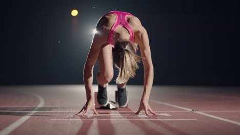 Fit-mixed-race-female-athlete-on-a-running-track-at-an-outdoor-sports-stadium-starting-a-race-from-starting-blocks-in-slow-motion-backlit-with-lens-flare.-Night